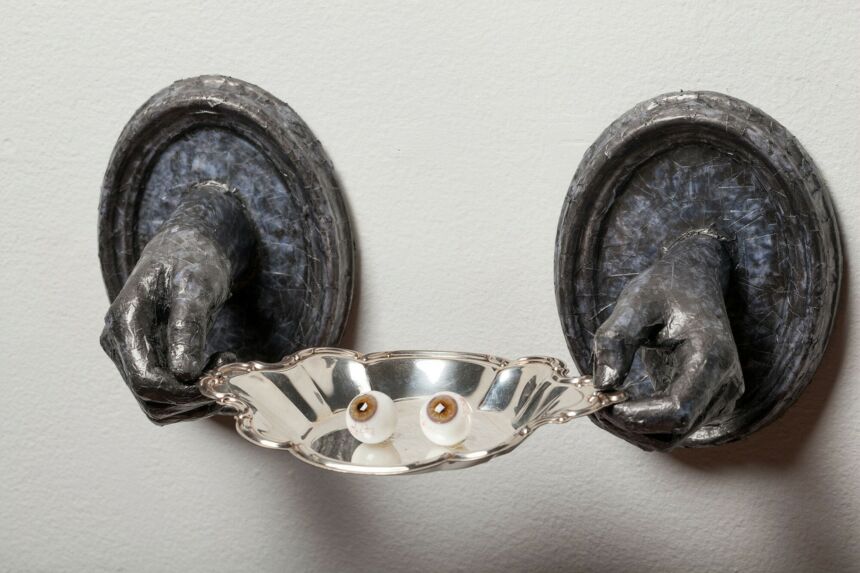 Close-up shot of the grey protruting hands holding a silver platter with two glass eyes.