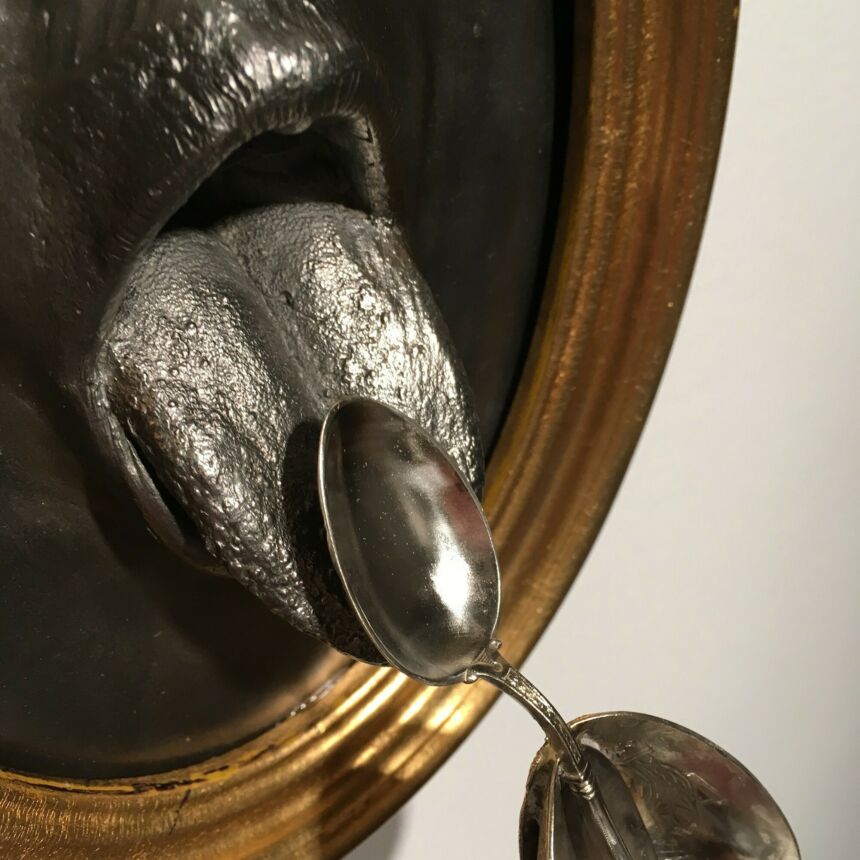 Detail shot of art installation of many silver spoons cascading from a painted-black disembodied mouth embedded in a wood frame.