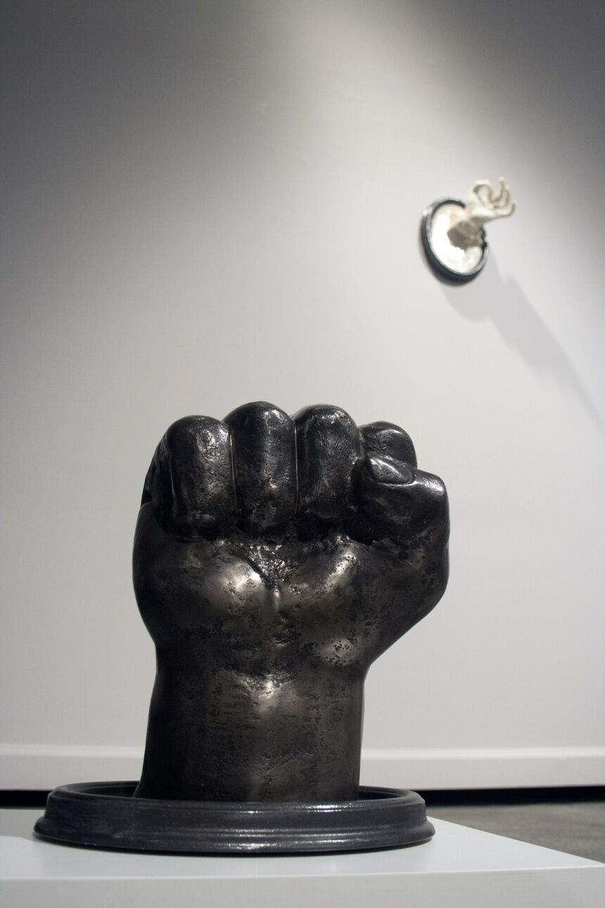 Large floor sculpture of black cast iron hand balled into a fist. In the background, a white feminine hand sculpture protrudes from a wood frame.