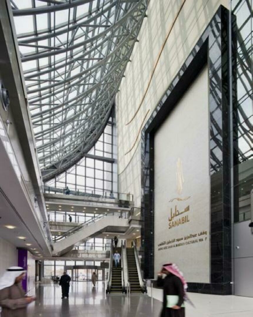Interior photo of a lobby with high vaulted glass ceilings, escalators in the distance, and visitors walking past.