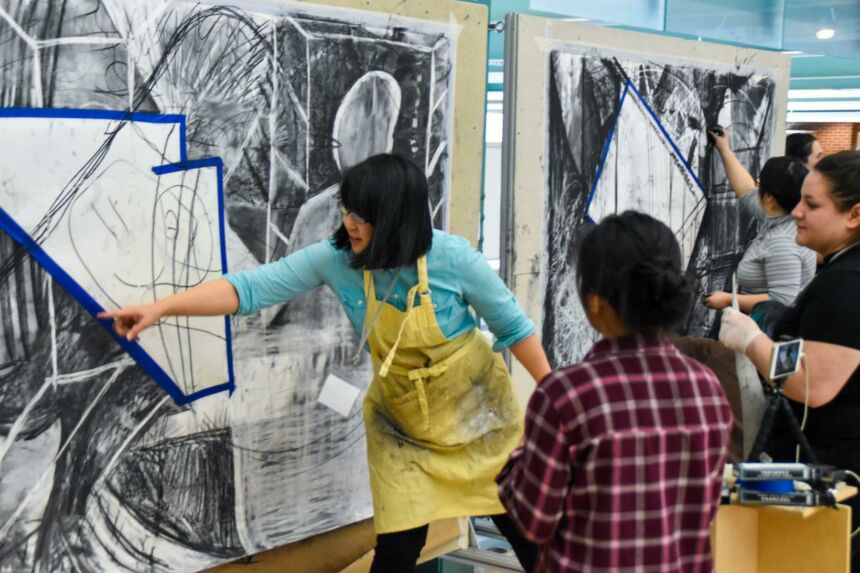 Photo of a woman pointing to a part of a large charcoal drawing while instructing two students.