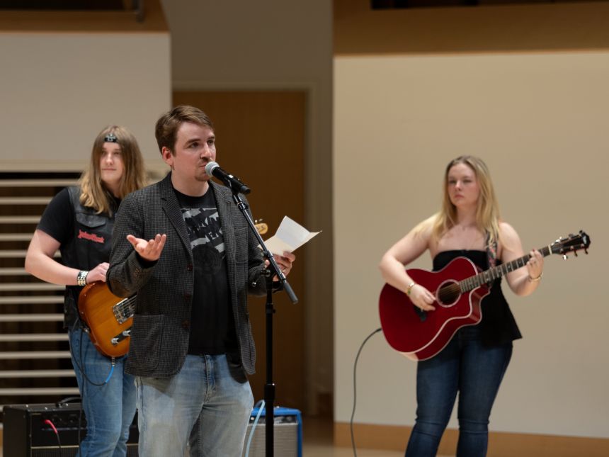 A young man in casual jeans, t-shirt, and unbuttoned overshirt speaking at a stand microphone on stage with two guitarist in the background.