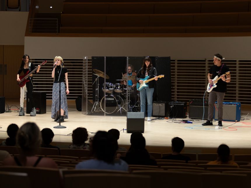 A 5-person rock band playing on stage in front of an audience at Penn State's Recital Hall.